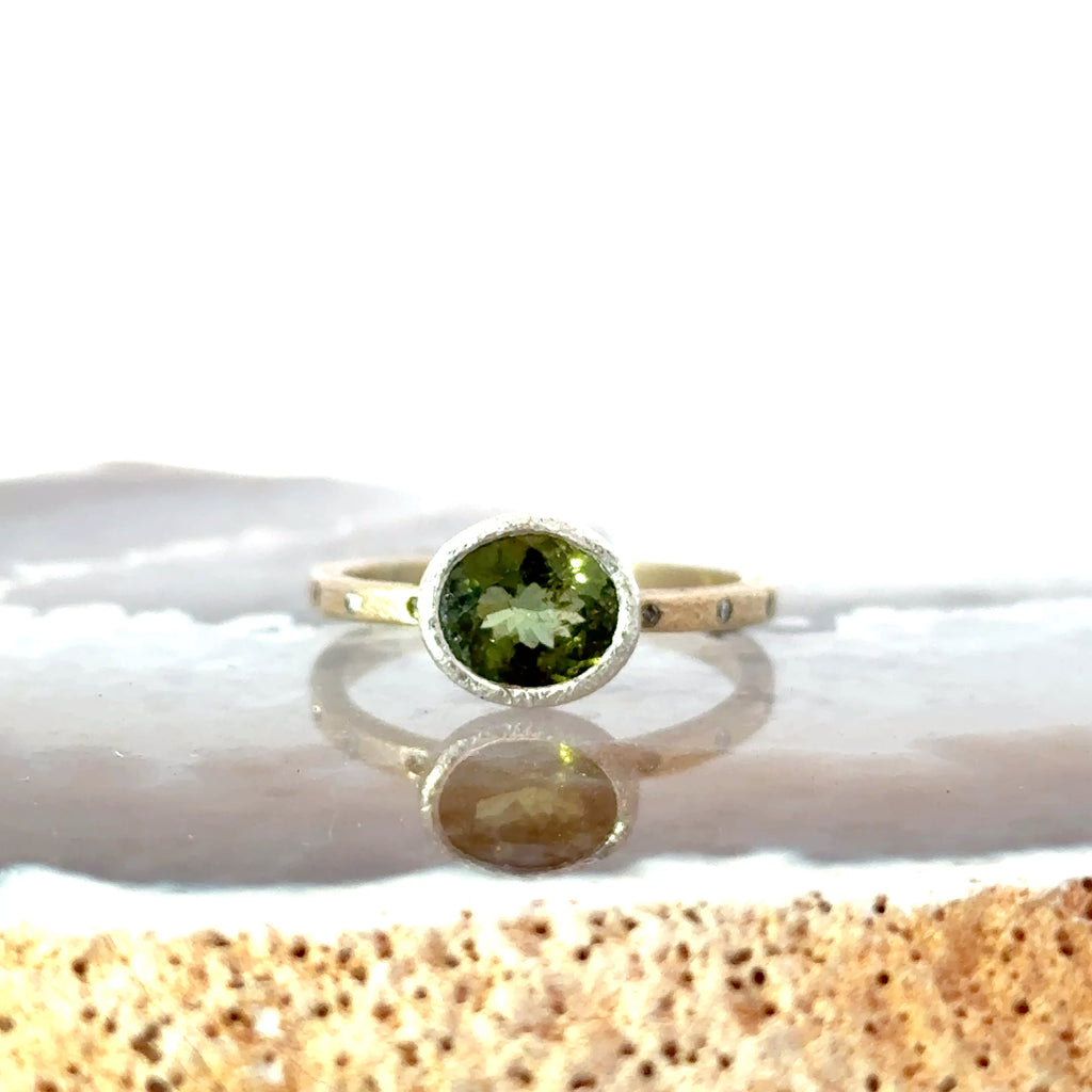 A silver ring with a round green gemstone, resting on a polished stone surface with a soft reflection. Champaign diamond accented ring band